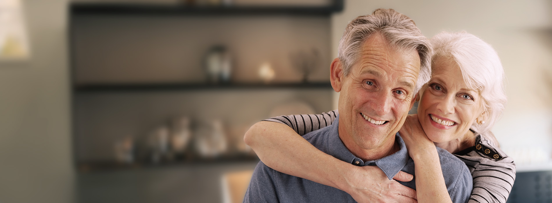 An elderly couple embracing warmly with smiles on their faces, set against a blurred background of a kitchen cabinetry.